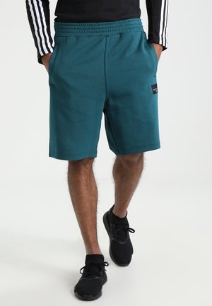 Mand iført blågrønne knælange shorts, sorte sneakers og en sort langærmet top med hvide striber på ærmerne, står med hænderne i lommerne.