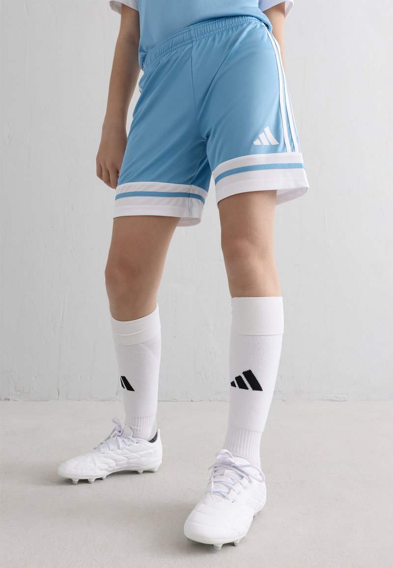 Person in hellblauen Sportshorts, weißen knielangen Socken und weißen Fußballschuhen, steht vor einer schlichten weißen Wand und einem weißen Boden.