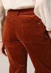 Pantalon en velours côtelé marron avec poches arrière, tissu texturé et design ajusté, avec des coutures visibles et un petit logo en métal.