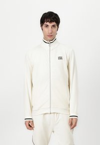 Weiße Zip-Jacke mit hohem Kragen, ribbeliger Textur und schwarzen Akzenten an den Bündchen und dem Reißverschluss. Logo auf der Brust.