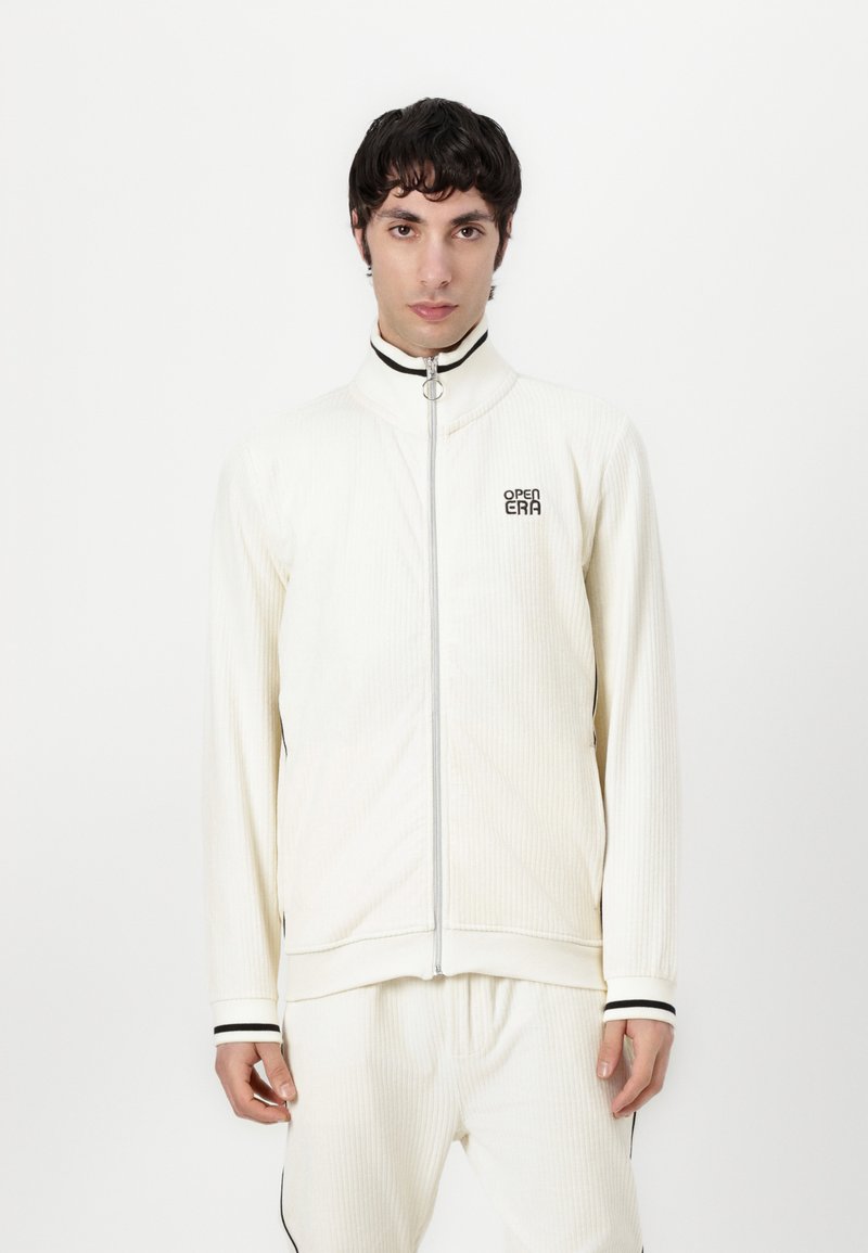 Veste blanche zippée avec un col haut, présentant une texture côtelée et des accents noirs aux poignets et sur la fermeture éclair. Logo sur la poitrine.