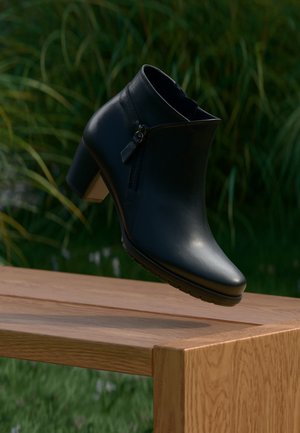 Schwarzer Stiefelette mit seitlichem Reißverschluss und Blockabsatz, schwebt über einem Holztisch im Freien mit grünen Pflanzen im Hintergrund.