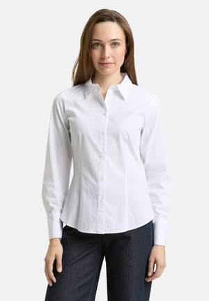 Woman wearing a fitted white button-up shirt with long sleeves and dark pants, standing against a plain light background.