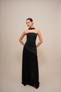 Woman in a strapless black evening gown with a sheer asymmetrical skirt panel, gold hoop earrings, and hair in a sleek bun, standing against beige background.
