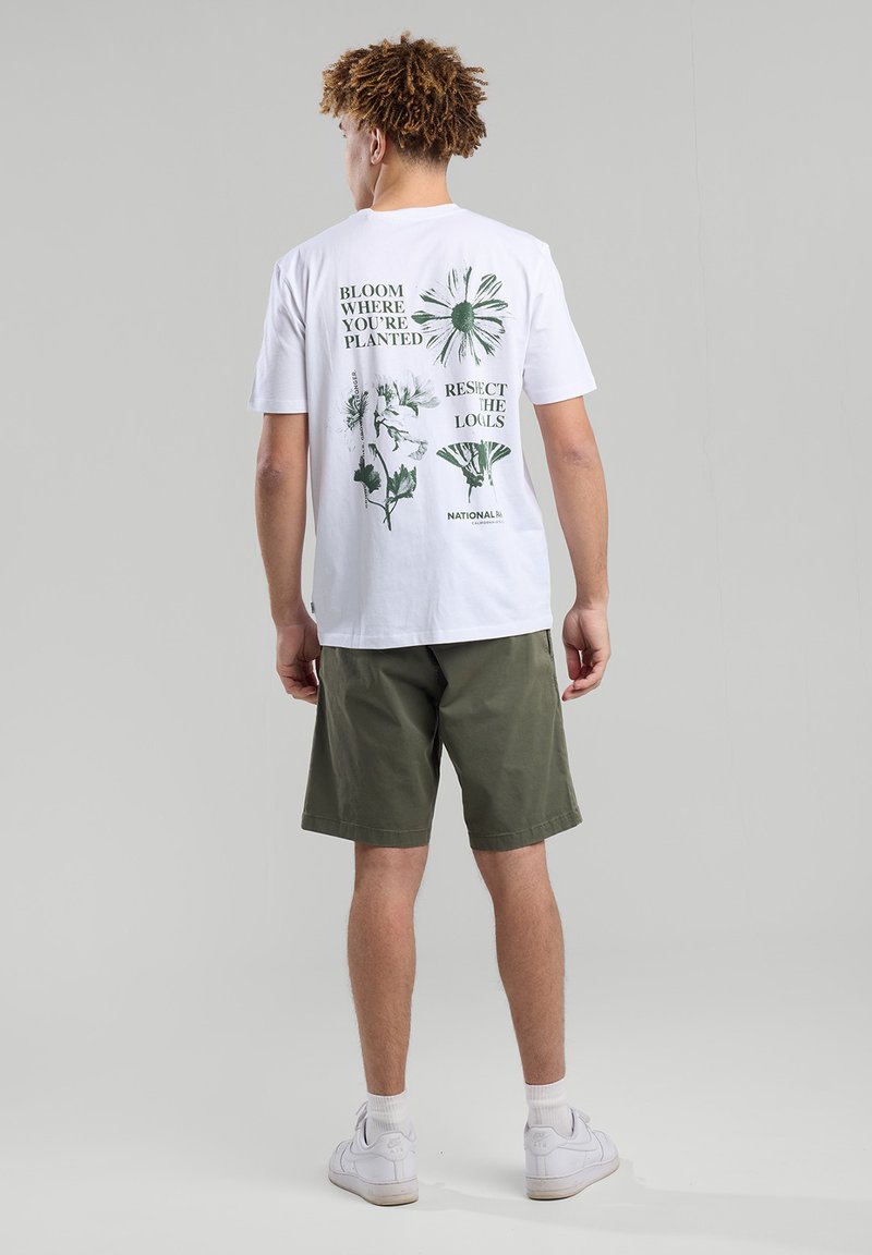 White t-shirt with green botanical prints and text on the back. Pair with olive green shorts. Model wearing white sneakers.