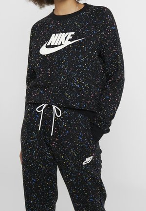 Osoba nosząca czarną bluzę i spodnie Nike z kolorowym wzorem smug farby oraz białymi logo Nike na klatce piersiowej i udzie.