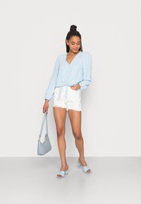 Ljusblå långärmad blus med knapprad och knytning i midjan, i kombination med vita slitna shorts, blå klacksandaler och handväska.