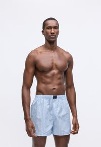 En atletisk mann med mørk hud som har på seg lyseblå stripete boxershorts og står foran en enkel hvit bakgrunn.