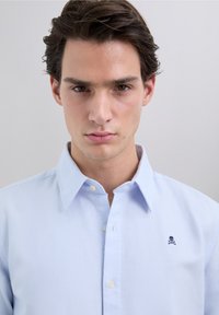 Camisa a rayas azul claro con cuello clásico, cierre de botones en la parte delantera y pequeño logo bordado de calavera con huesos cruzados en el pecho.
