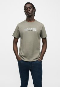 Άνδρας με πλεξούδες, που φοράει μια ελιά πράσινη μπλούζα Calvin Klein και σκούρο μπλε τζιν, με τα χέρια στις τσέπες, στέκεται μπροστά από μονόχρωμο φόντο.