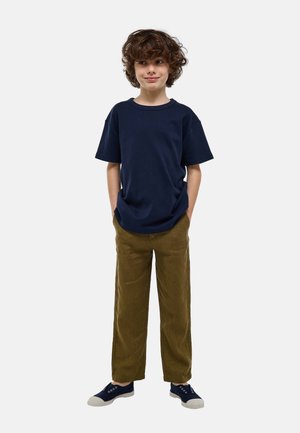 Enfant aux cheveux bouclés portant un t-shirt bleu marine, un pantalon vert olive et des baskets bleu marine, debout les mains dans les poches, regardant sur le côté.