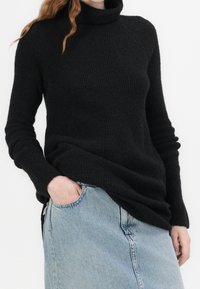 Personne portant un pull noir côtelé à col roulé avec manches longues et un jean en denim bleu clair taille haute, une main dans la poche.