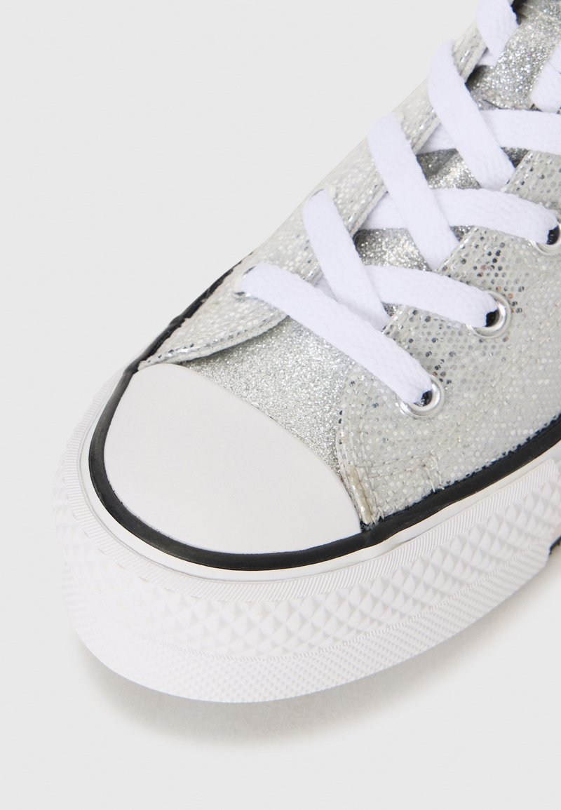 Zapatilla de lona plateada con purpurina, suela de goma blanca, detalles en negro y cordones blancos. Punta redondeada con patrón de tracción texturizado.