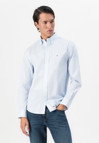 Mann i lyseblåmønstret skjorte med knapp og blå jeans, står med den ene hånden i lommen og ser mot venstre mot hvit bakgrunn.
