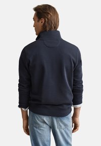 Felpa blu navy con collo alto e polsini a coste, con dettaglio circolare sulla schiena, indossata con jeans azzurri.