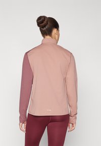 Langermet treningsgenser i mauve og burgunder fargeblokkering, laget av mykt, elastisk stoff med krage og logo på kanten.