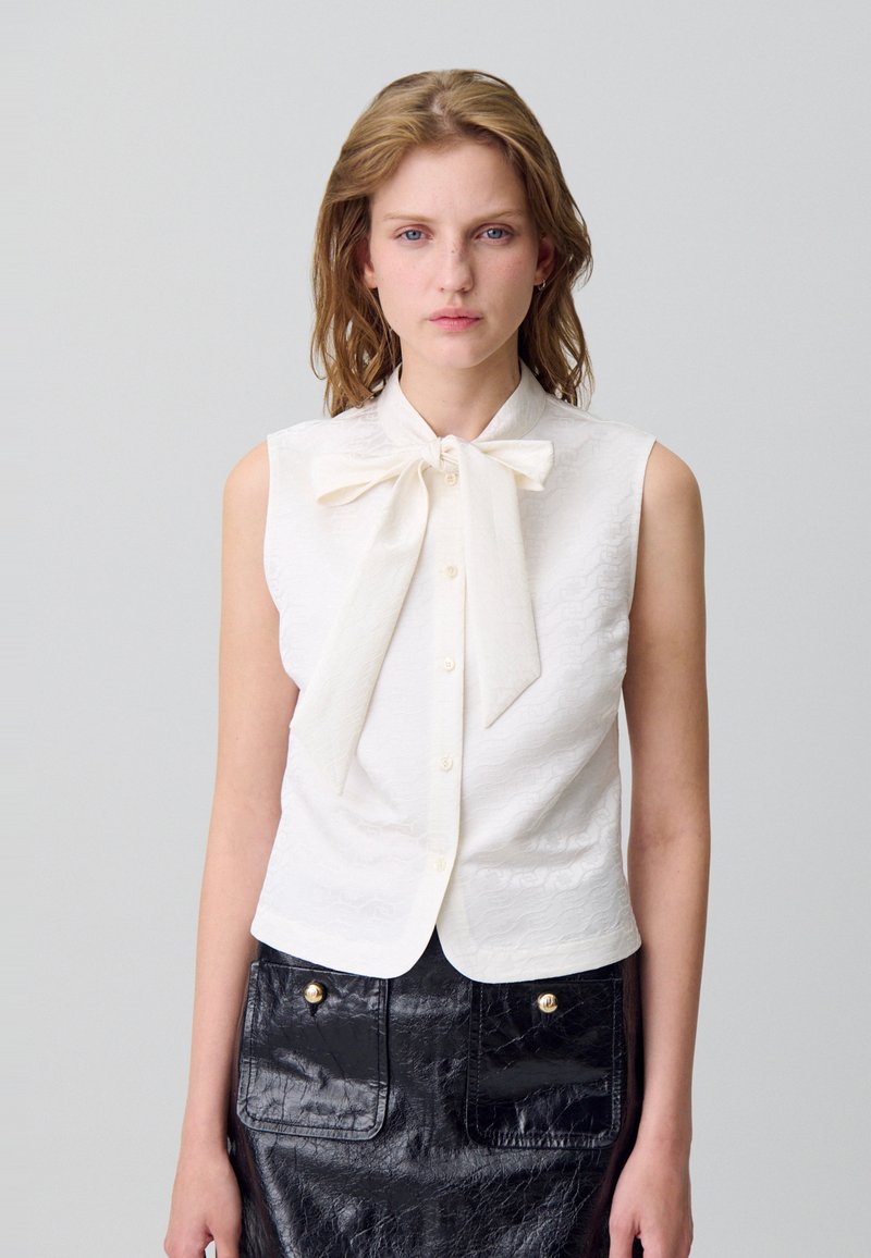 Blusa blanca sin mangas con un lazo en el cuello, que presenta un acabado texturizado. Combinada con una falda de cuero negro brillante de cintura alta.