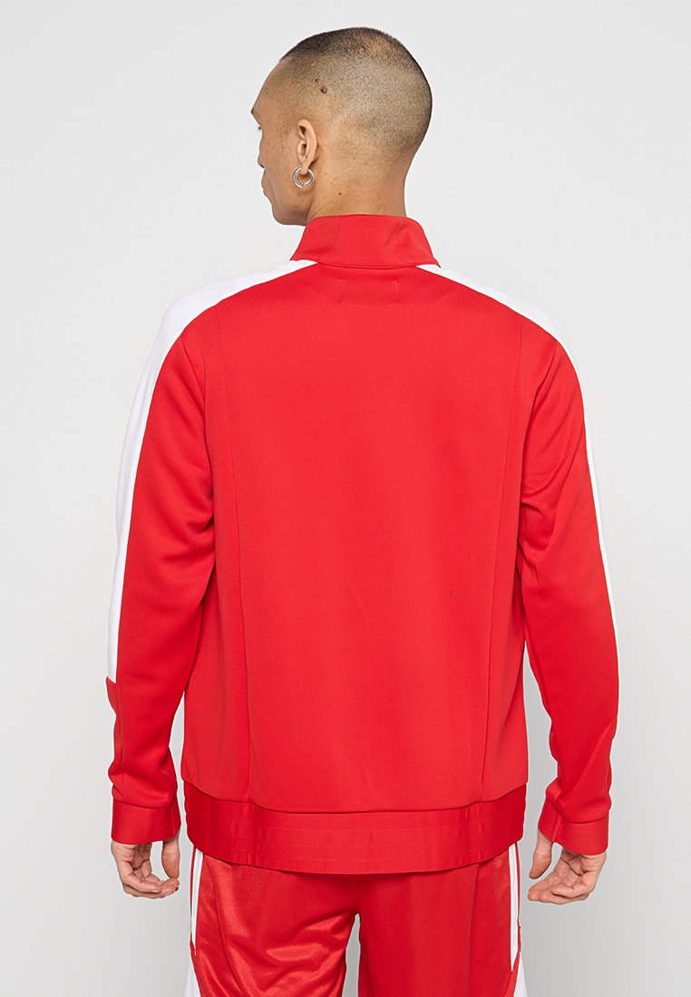 Chaqueta atlética roja con un alto cuello, mangas largas y paneles laterales en blanco contrastante. Fabricada con un material liso y ligero.