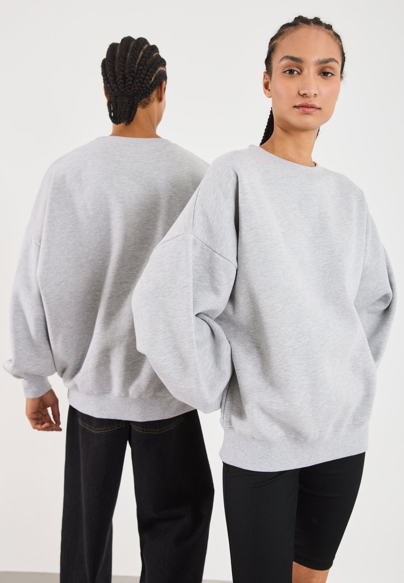 Felpa oversized grigia chiara con spalle scese e polsini a coste, abbinata a pantaloni neri. Tessuto morbido, vestibilità rilassata.