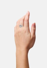Anello in argento con un design floreale e dettagli in openwork, appoggiato su una mano. Texture liscia, finitura lucida e forma arrotondata.