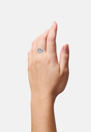 Silberner Ring mit einem floralen Design und offenen Akzenten, auf einer Hand präsentiert. Glatte Textur, polierte Oberfläche und runde Form.