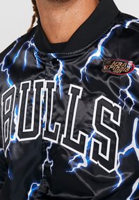 Musta satiininen bomber-takki, jossa näkyvillä rohkeat valkoiset "BULLS"-kirjaimet, siniset salamaniskugrafiikat ja kirjailtu NBA-finaalipatchi.