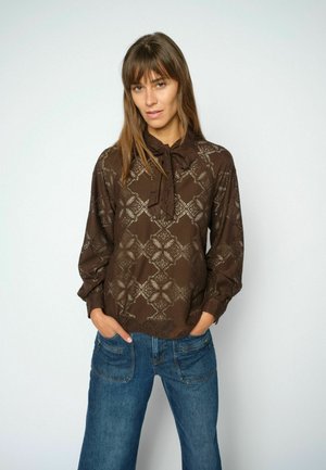 Woman wearing a brown patterned long-sleeve blouse with a neck tie and blue jeans, standing with hands in pockets against a plain background.