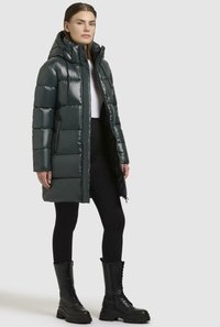 Polstret, blank mørkegrøn frakke med hætte, der har en frontlynlås, sidelommer og en knælang design, båret over sorte leggings og støvler.