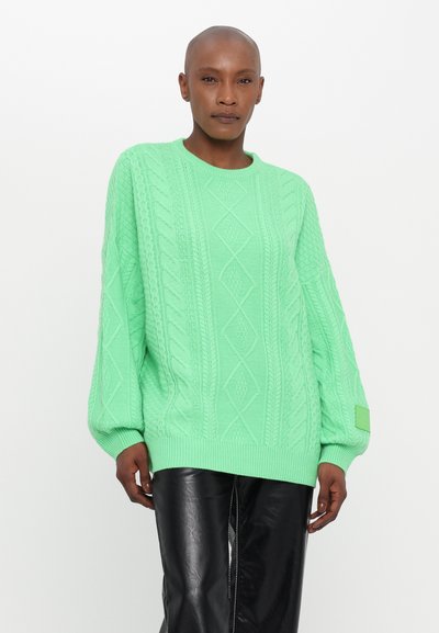 Maglione a maglia verde brillante con motivi a treccia e diamante, polsini a coste e una toppa decorativa sulla manica. Tessuto morbido, vestibilità rilassata.