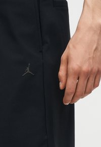 Κοντινή λήψη ενός χεριού που ακουμπά δίπλα σε ένα μαύρο ένδυμα με ένα μικρό, σκοτεινό λογότυπο Jordan Jumpman κοντά στη ραφή της τσέπης.