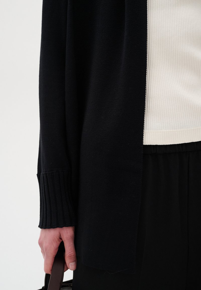 Langer schwarzer Cardigan mit gerippten Ärmeln, getragen über einem cremefarbenen ribbed Top. Eine Hand hält eine braune Tasche, schwarze Hosen sind teilweise sichtbar.