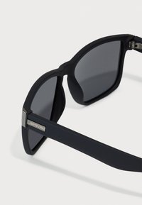 Ochelari de soare negri cu finisaj mat, formă rectangulară, lentile întunecate și un accent argintiu pe tâmplă cu branding "Santa Cruz".