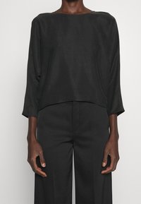 Blouse noire en tissu lisse, avec une coupe décontractée, un col bateau et des manches trois-quarts, assortie à un pantalon noir.