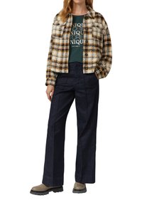 Geruite wolmix jas in crème, bruin en mosterdtinten over een groene graphic t-shirt, gecombineerd met donkerblauwe wijde jeans en beige schoenen.