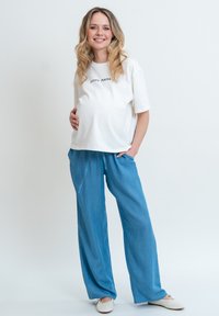 Těhotenský outfit skládající se z bílé bavlněné trička s potiskem "COOL MAMA" a volných modrých kalhot v jemném materiálu, doplněný o světlou obuv.