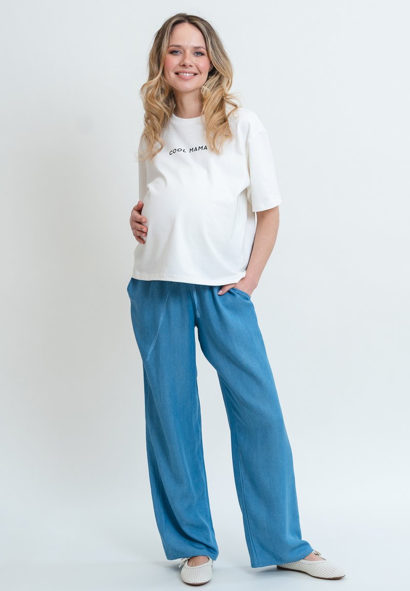 Těhotenský outfit skládající se z bílé bavlněné trička s potiskem "COOL MAMA" a volných modrých kalhot v jemném materiálu, doplněný o světlou obuv.