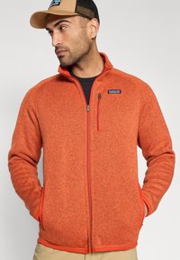 Πορτοκαλί ζακέτα φλις με φερμουάρ στο μπροστινό μέρος, ψηλό γιακά και κόκκινες αντιθέσεις. Διαθέτει τσέπη στο στήθος και υφή στο ύφασμα.