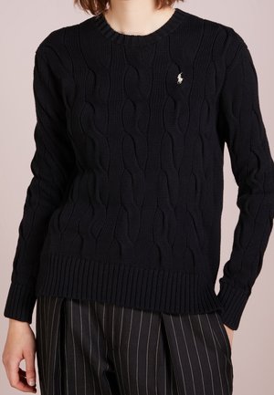 Person trägt einen schwarzen, kabelgestrickten Pullover mit gerippten Bündchen und Saum, kombiniert mit schwarzen Hosen mit Nadelstreifen.