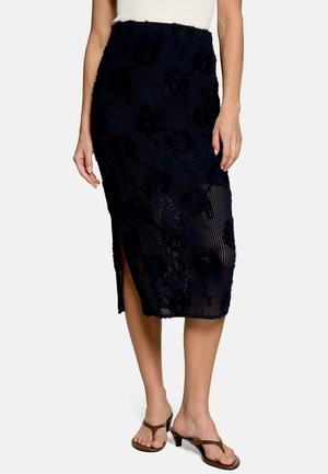 Kobieta w czarnej teksturowanej spódnicy midi z kwiatowym wzorem i bocznym rozcięciem, w połączeniu z brązowymi sandałami na obcasie i białą bluzką bez rękawów.