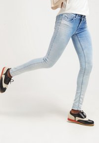 Jeans skinny de color azul claro con un lavado desvaído y diseño de cinco bolsillos, combinados con zapatos de vestir negros y crema con detalles de punta de ala.