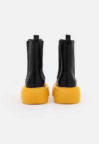 Botas de tobillo de cuero negro con un caña alta, que presentan una gruesa suela amarilla texturizada. Incluyen tiradores para facilitar el calzado.