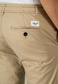 Pantalones de algodón beige con un bolsillo trasero visible, cierre de botón negro y una etiqueta blanca cosida. Textura suave y diseño casual.