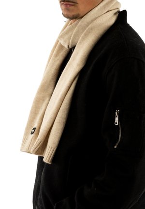 Uomo con giacca nera dotata di tasca con zip sulla manica e sciarpa di lana beige avvolta attorno al collo, labbra e mento visibili.