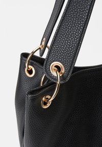Black textured handbag featuring gold-tone hardware and circular grommets. The shoulder straps are flat and seamlessly attached.