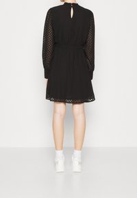 Femme portant une robe noire texturée longueur genou avec de longues manches transparentes, des chaussettes blanches jusqu'à la cheville et des baskets blanches ornées, vue de dos.