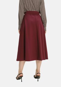 Bordeaux Midi-Rock aus glattem Stoff mit ausgestellter Silhouette, ausgestattet mit einem Bund und einem dezenten Nahtdetail auf der Rückseite. Getragen mit schwarzen High Heels.