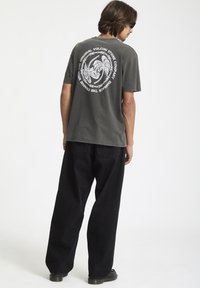 Mørkegrå bomuldst-shirt med hvid grafisk design på ryggen, parret med løse sorte jeans og sorte fodtøj.