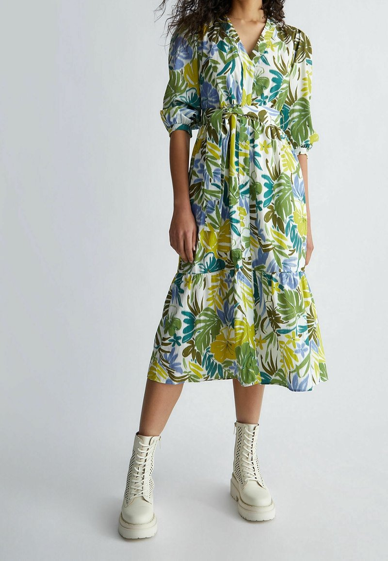Vestido midi floral con patrones tropicales en verde, azul y amarillo, con cuello en V, mangas abullonadas y cinturón en la cintura; combinado con botas blancas hasta el tobillo.