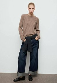 Brązowy sweter z prążkowanego materiału, ciemnoniebieskie szerokie dżinsy z podwiniętymi nogawkami, ciemny sweter związany w talii, czarne mokasyny, minimalistyczne tło.