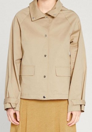 Veste beige à boutons avec col, poches à rabat et sangles ajustables aux poignets portée sur une jupe couleur moutarde.